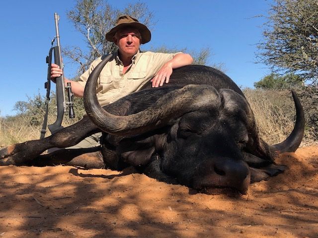 Man in hat poses with a dead buffalo and rifle in a sunny, sandy field.