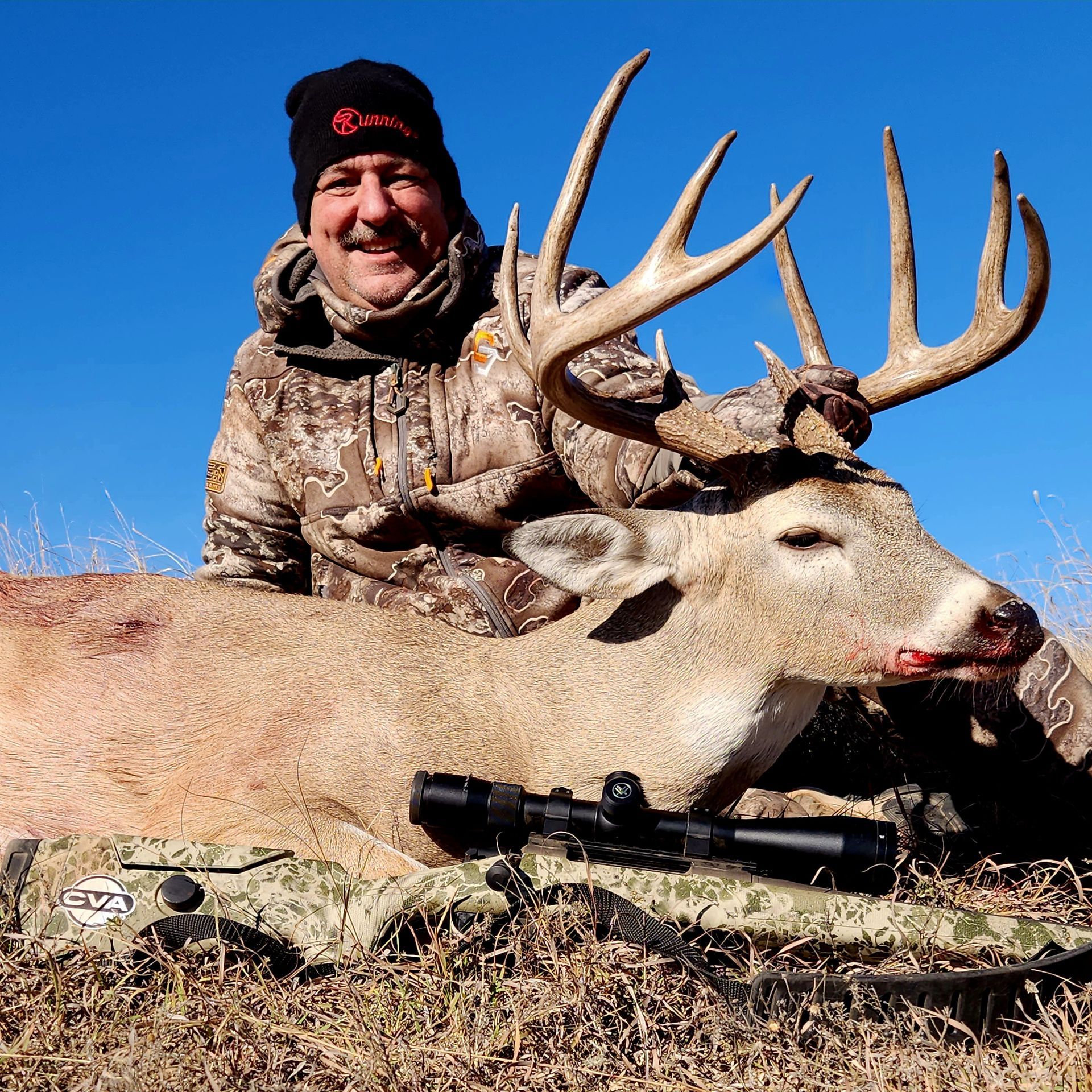 Hunter smiles, posing with a large buck. The deer lies in tall grass, rifle in front. Clear, blue sky.
