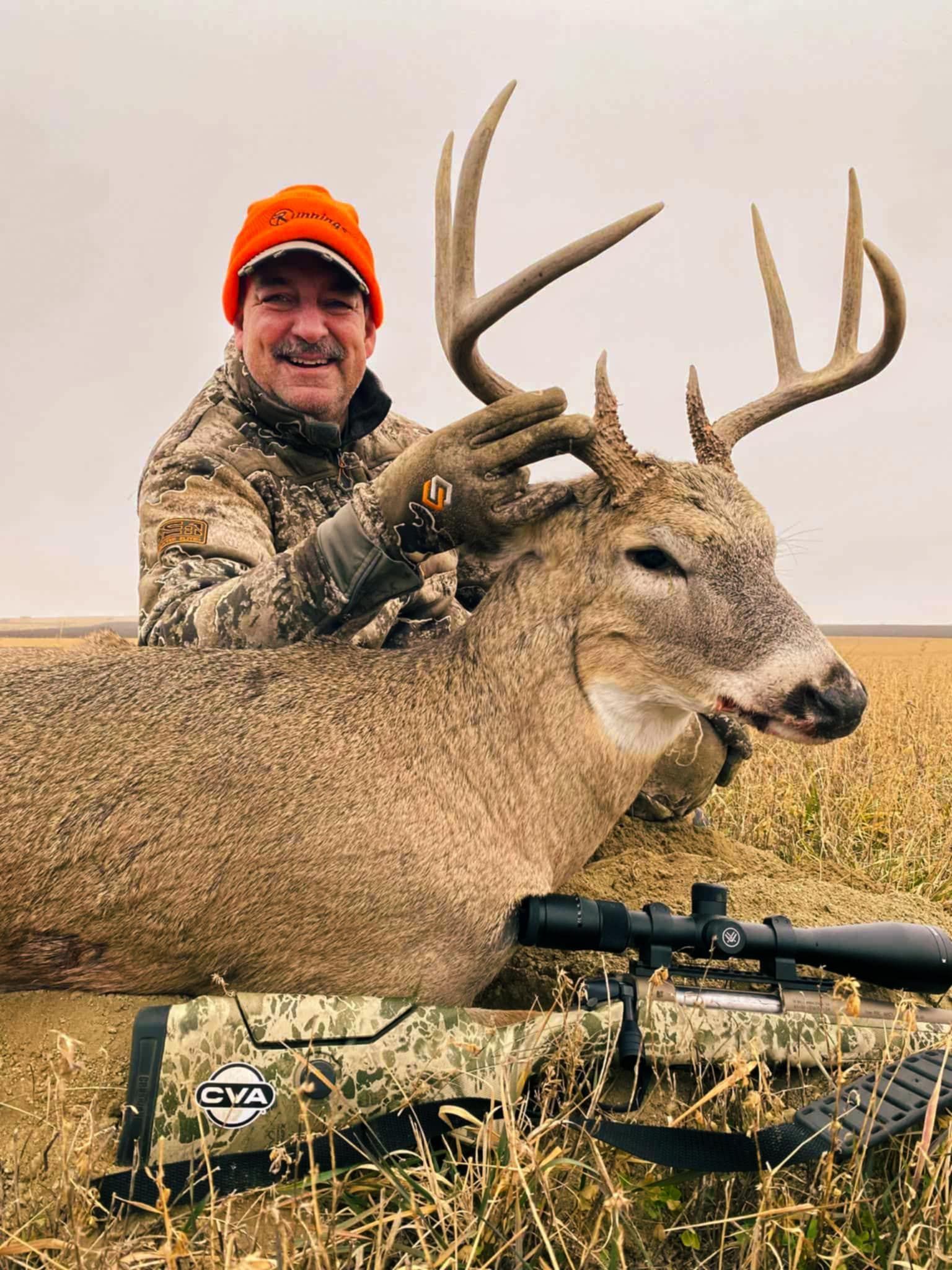 Man in orange hat and camo jacket smiles, holding antlers of a deer in field; rifle in foreground.