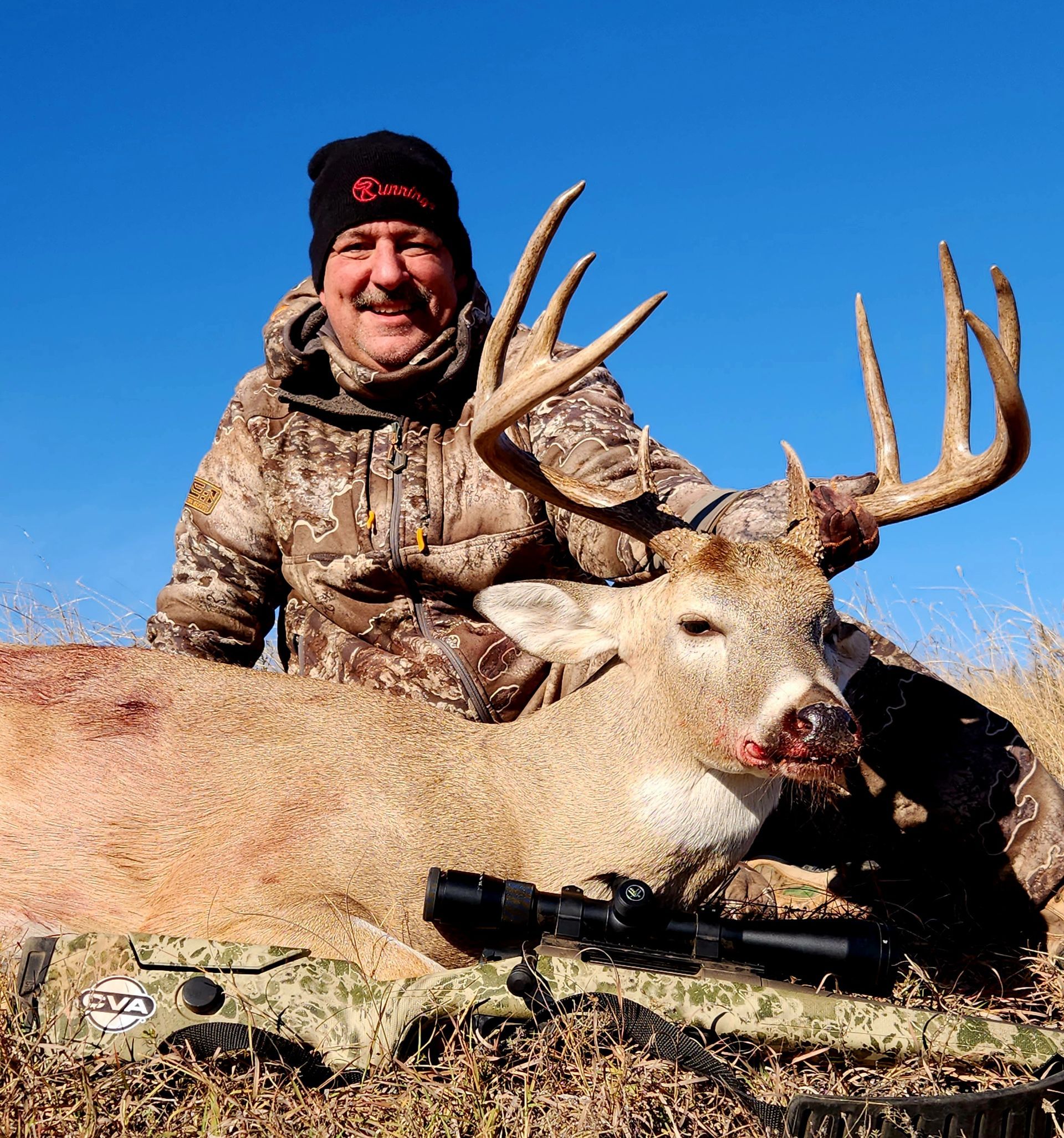 Man in camo with a large deer, posed outdoors under a blue sky, smiling at the camera.