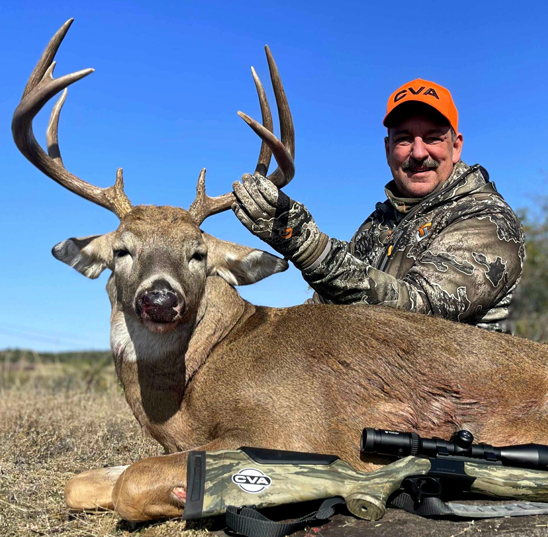 Man in camouflage and orange cap with a large buck. Rifle is next to the deer. Blue sky, outdoor setting.