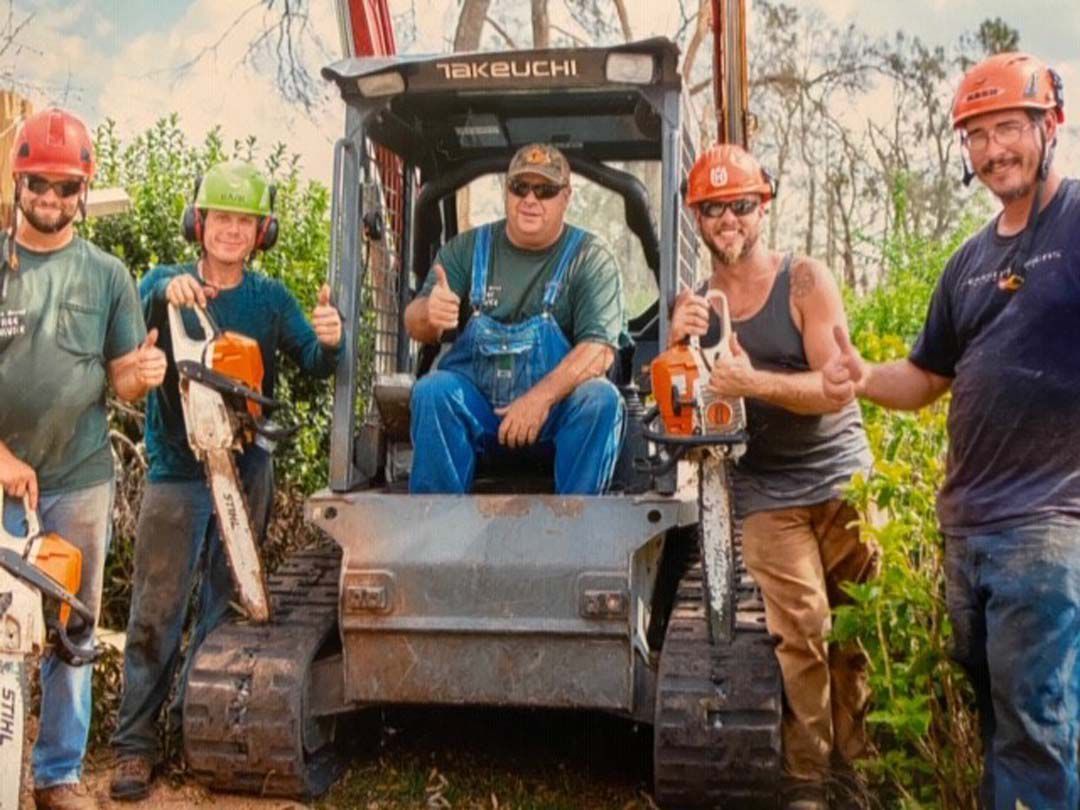 Five people with chainsaws and a skid steer, giving thumbs up in a wooded area.
