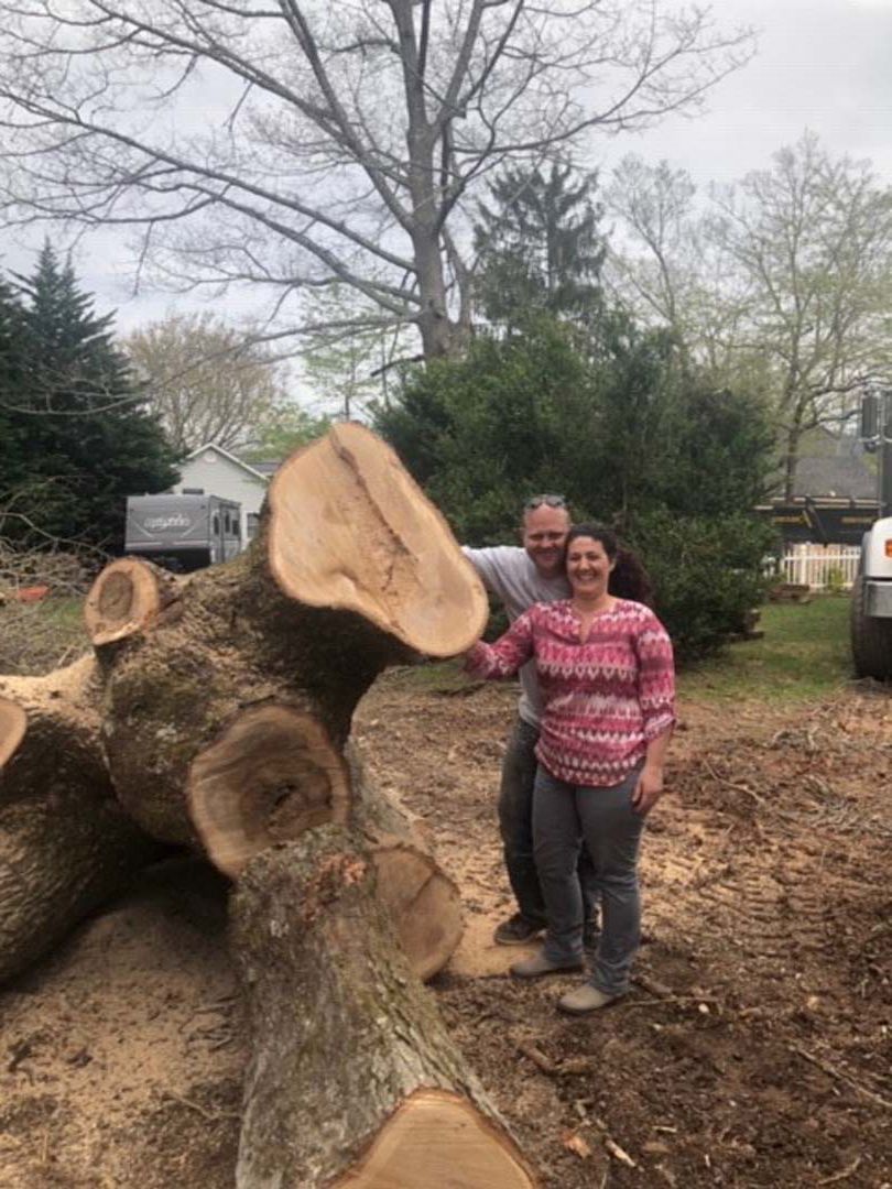 Couple stands next to a felled tree trunk, smiling. Outdoors, brown and green, cloudy sky.