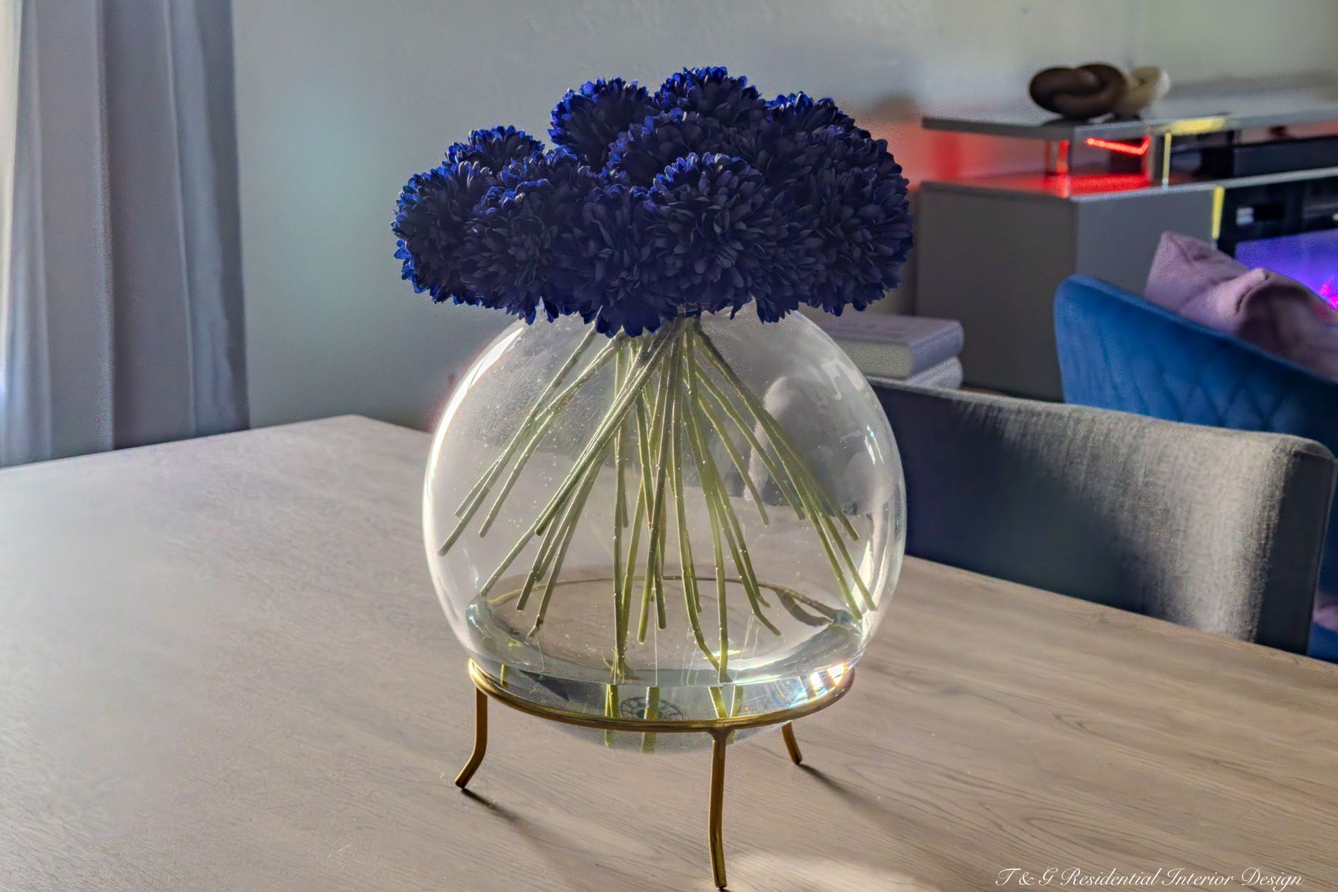 Blue flowers in a round glass vase on a gold stand, sitting on a light-colored table.