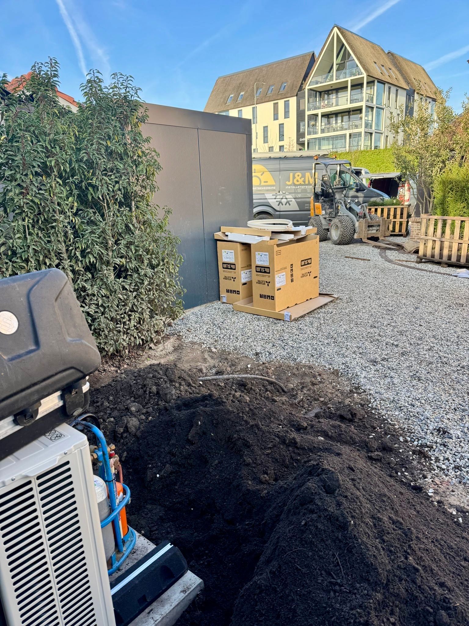 Bouwplaats met grind, dozen, trilplaat en een gebouw op de achtergrond onder een blauwe hemel.