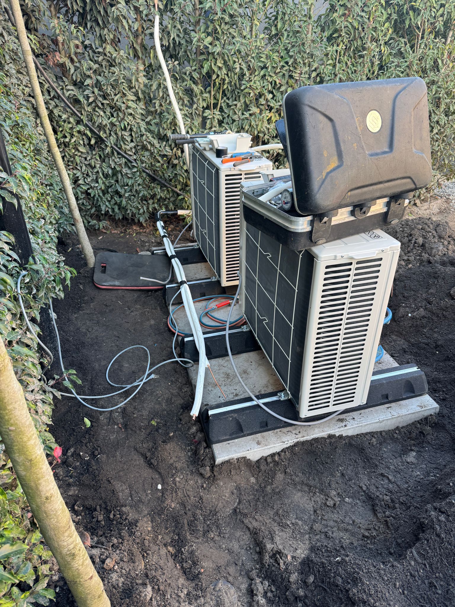 Twee buitenunits voor de airconditioning staan ​​op een grindfundering naast een heg en een boomstam.