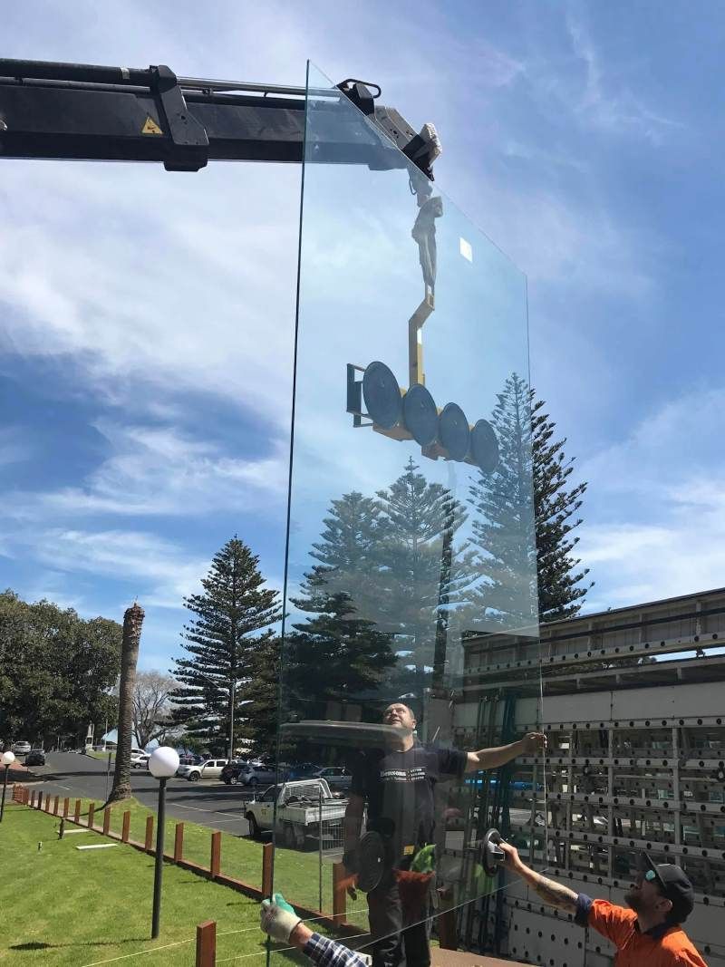 Crane lifting a large glass panel outdoors near workers and a building under a blue sky