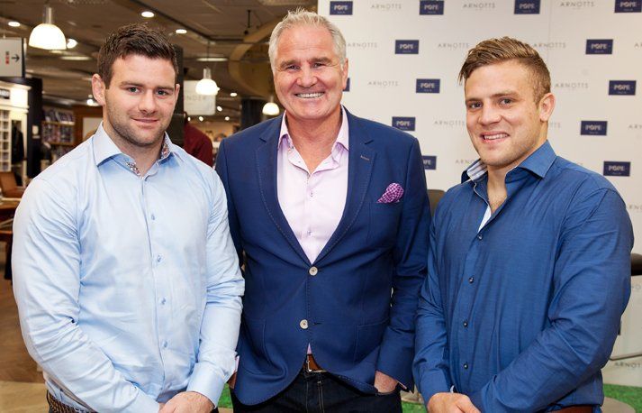 Three men in dress shirts and jackets pose indoors. Two men wear blue shirts, and one has a pink shirt.