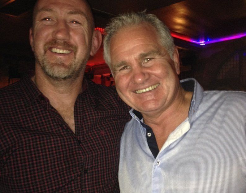 Two men smiling, one in a checked shirt, the other in blue, in a dimly lit bar setting.