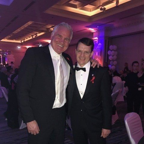 Two men in formal attire, smiling, with arms around each other in a decorated ballroom.