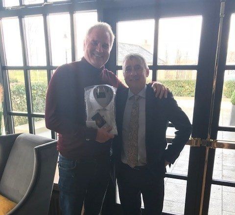 Two men posing; one holds a shirt in plastic; they stand by windows.