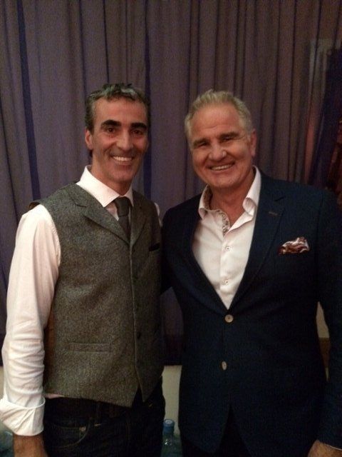 Two men smiling, posing together. One wears a vest, the other a navy suit. Inside setting.