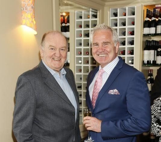 Two men smiling, one in a blue blazer with a drink, the other in a gray jacket. Wine bottles in the background.