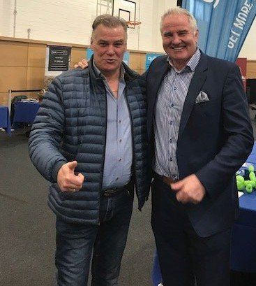 Two men smiling, posing. One in blue jacket, other in suit jacket, indoors near display tables.
