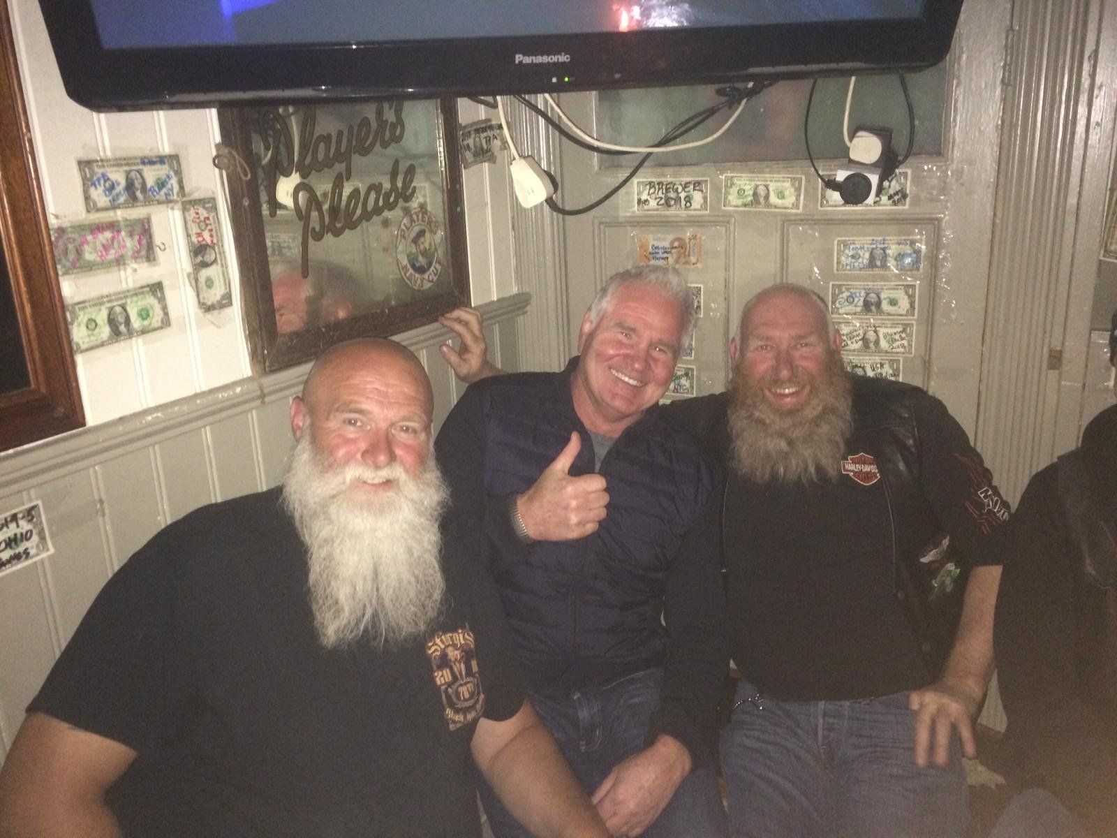 Three men in a bar. Two have long white beards. Man in the middle gives a thumbs-up.