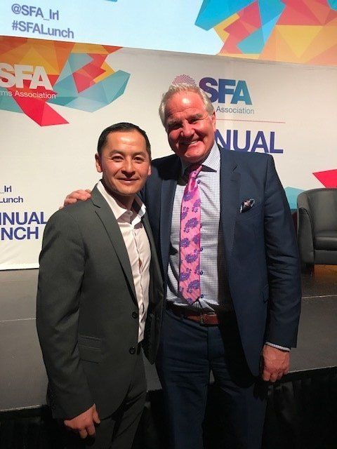 Two men pose for a photo at an event; one in a blazer, the other in a suit. They are smiling.