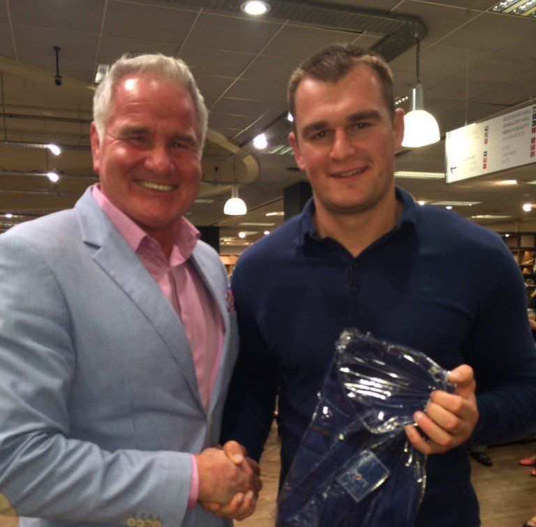 Two men shaking hands in a bookstore. One wears a blue blazer, the other a navy shirt and holds a package.