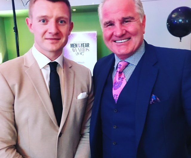 Two men in suits posing for a photo. The man on the left wears tan and black tie. The other wears navy, a pink patterned tie.