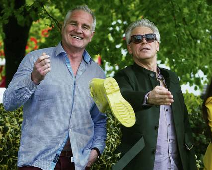 Two men outdoors, one holding a yellow boot, the other pointing; sunny day.