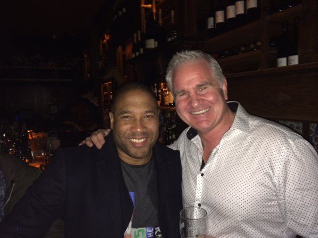 Two men smiling at a bar. One has arm around the other's shoulder, dark setting.