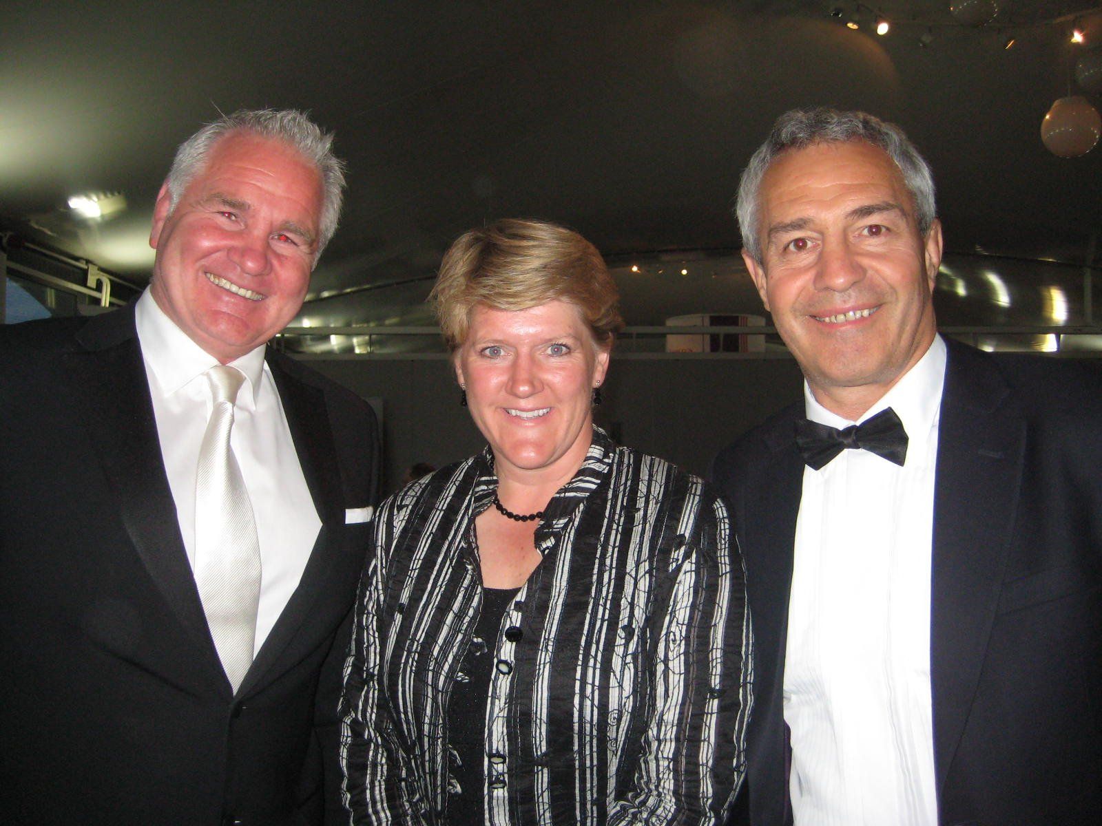 Three people smiling together in formal wear at an event; dark background.