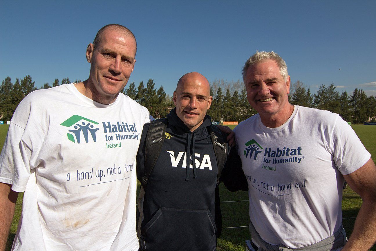 Three men wearing Habitat for Humanity shirts pose outdoors on a sunny day.