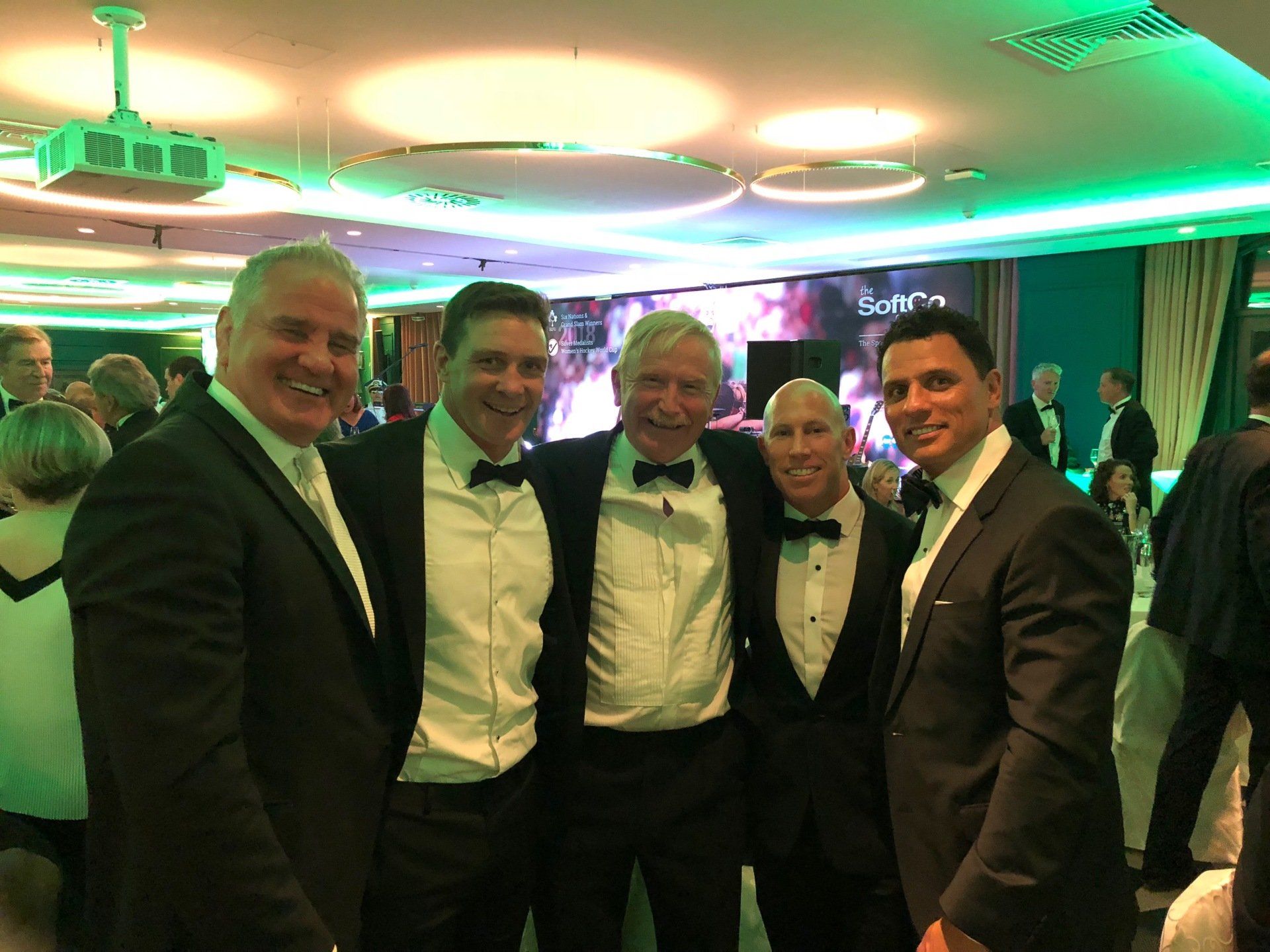 Five men in tuxedos pose for a photo at a gala. Brightly lit room with background screen.