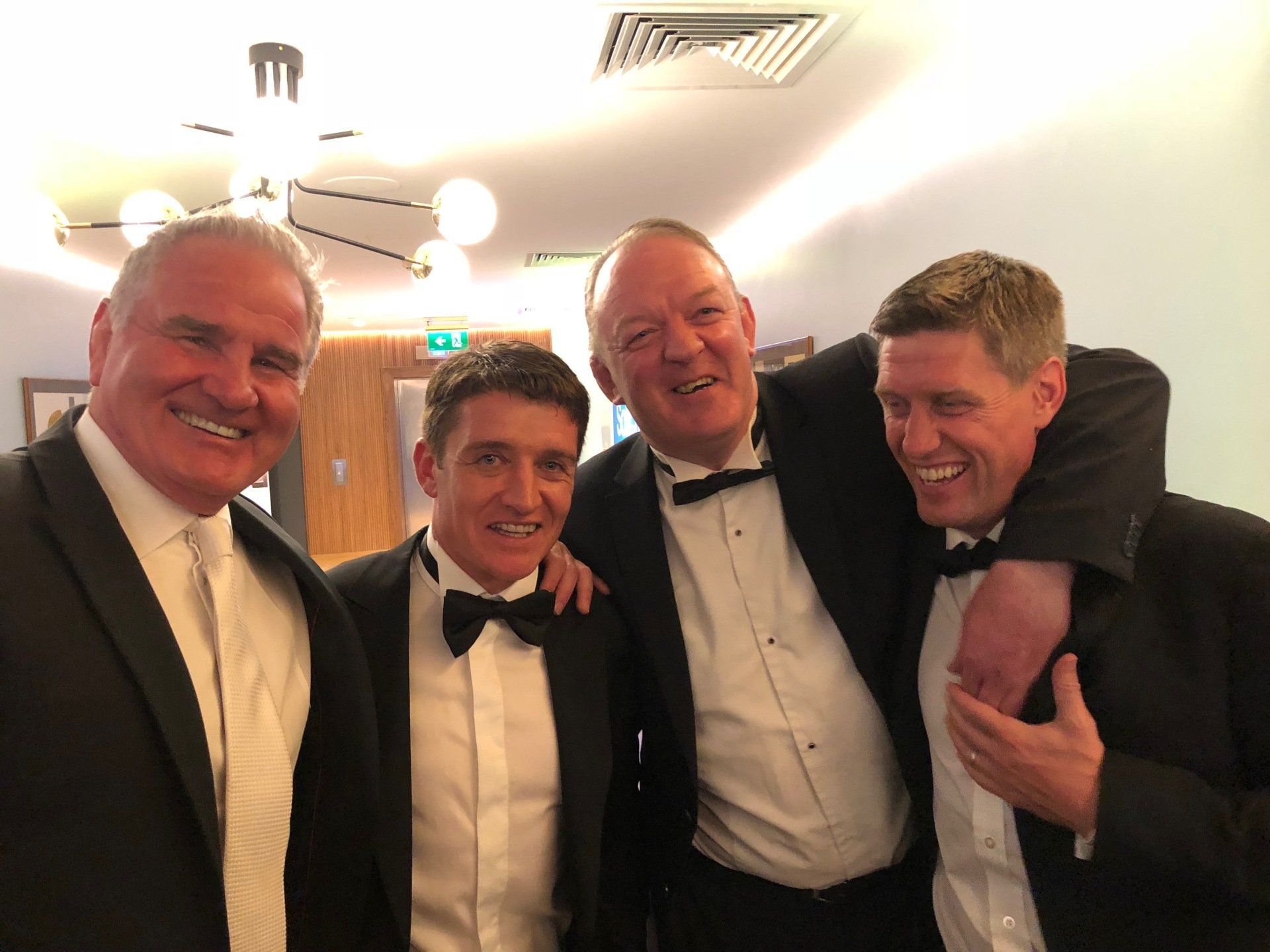 Four men in tuxedos smile and pose together in a brightly lit room.