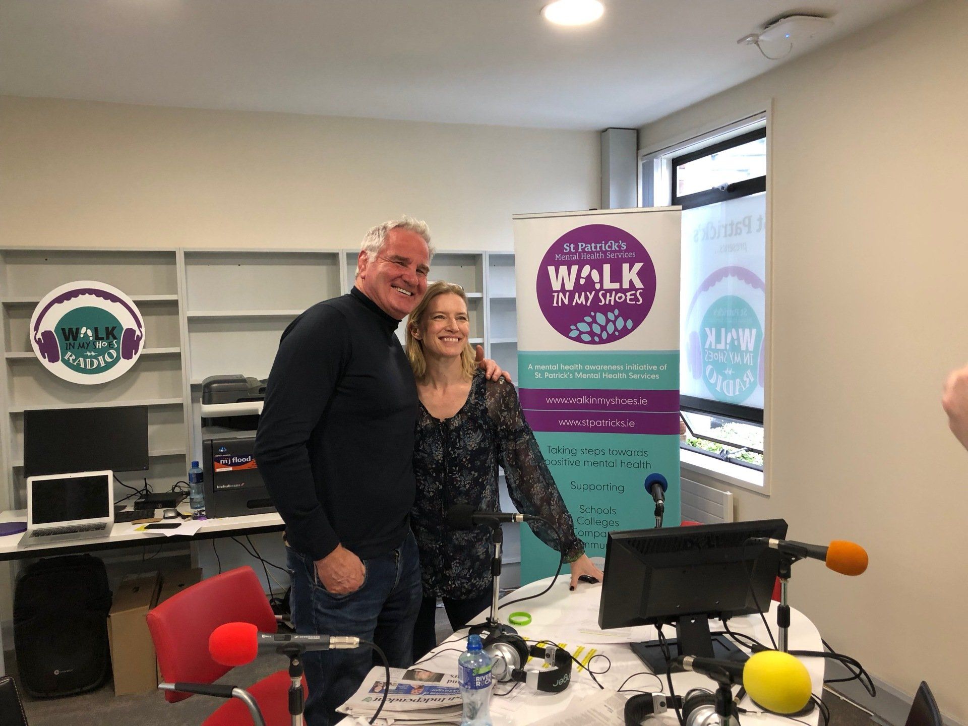 Two people pose in a radio studio. A man in a black sweater and woman with floral top smile near a 