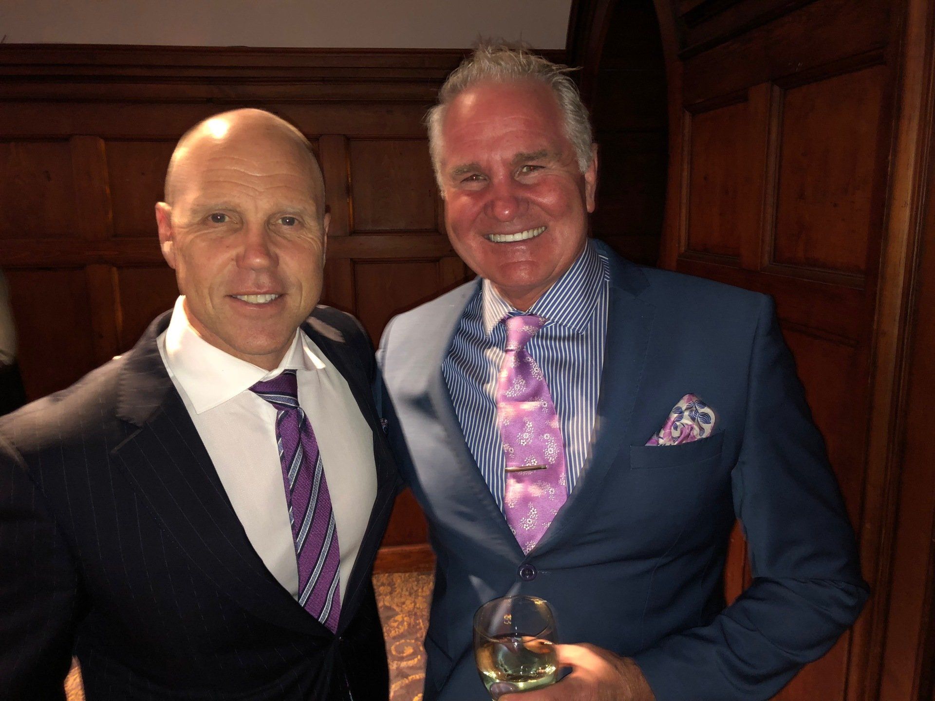 Two men in suits smile, posing indoors. One wears a blue suit, the other, black. Both have patterned ties.