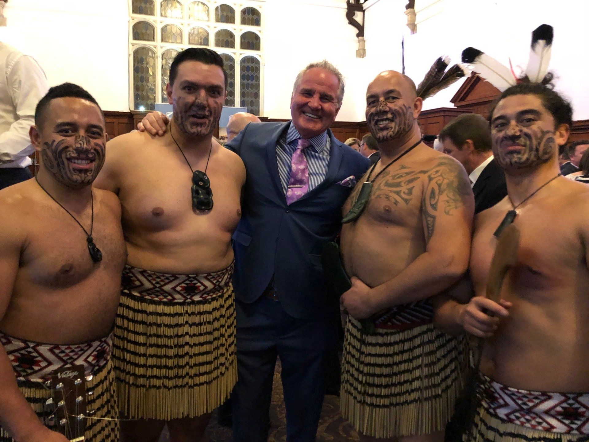 Man in suit poses with five men wearing traditional Maori attire at an event.