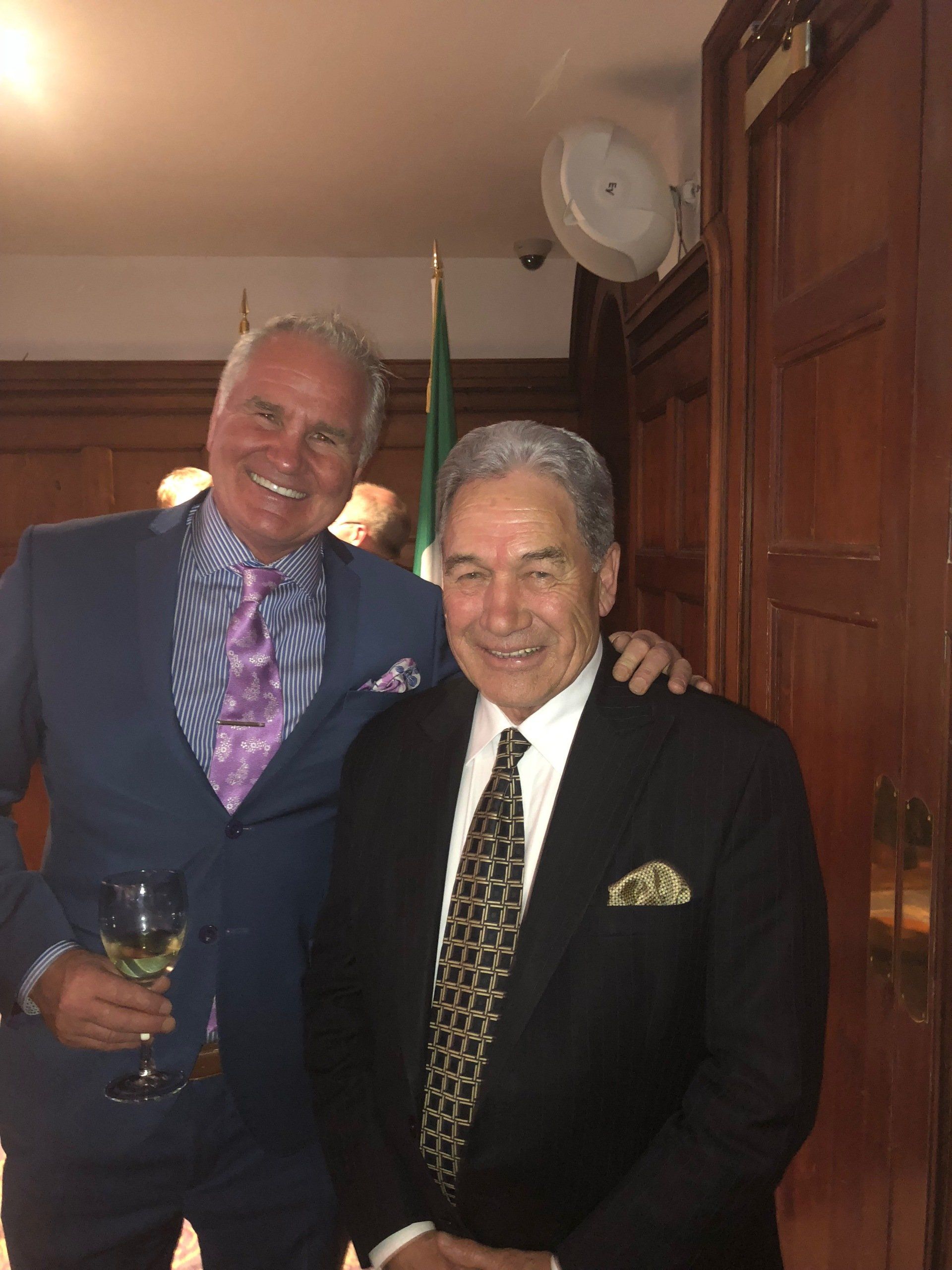 Two men in suits pose, one with arm around the other's shoulder, smiling. Indoors, near a flag.