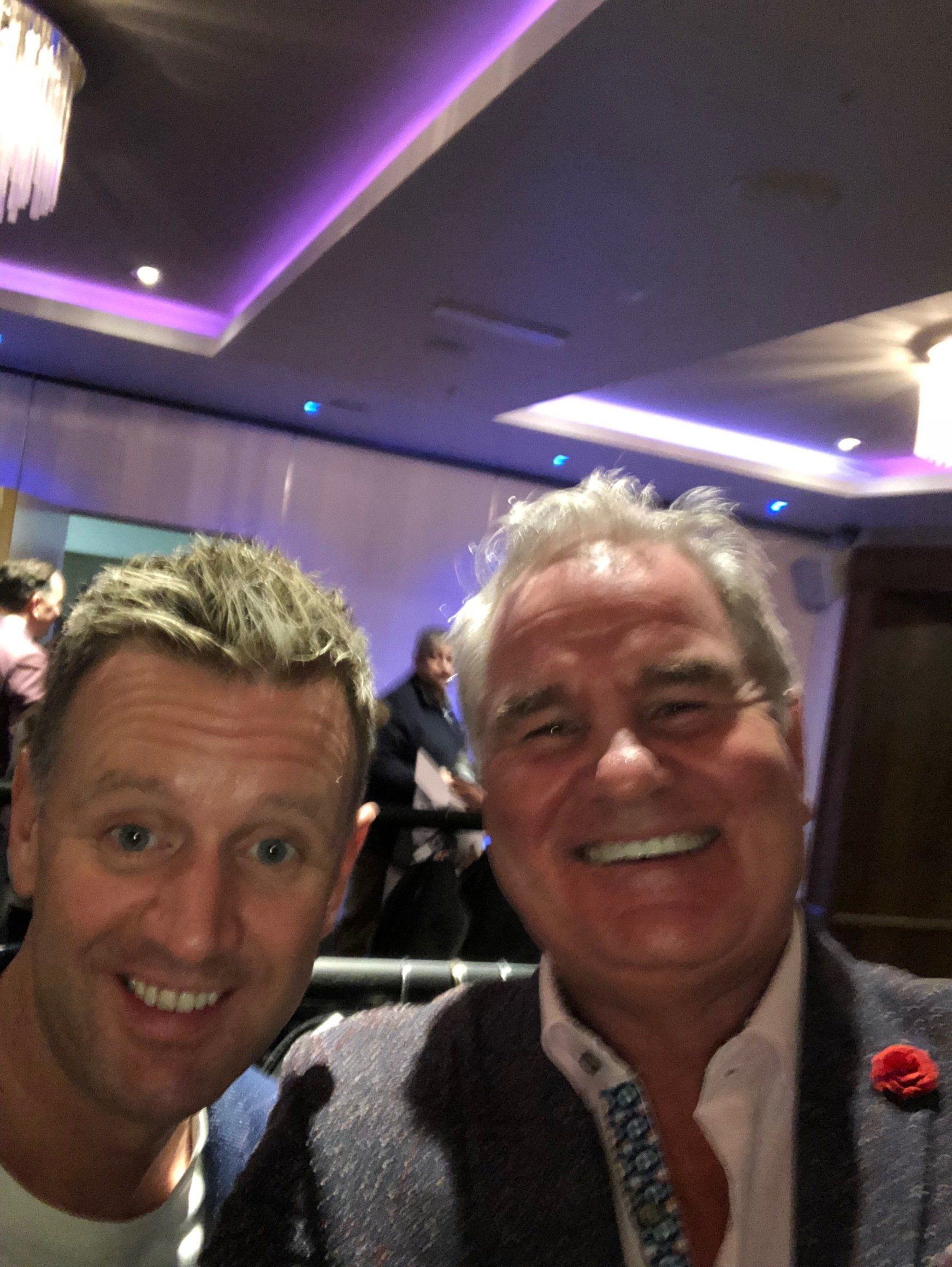 Two smiling men taking a selfie indoors. One wears a blazer with a red flower, the other a plain shirt. Lit room, purple lights.