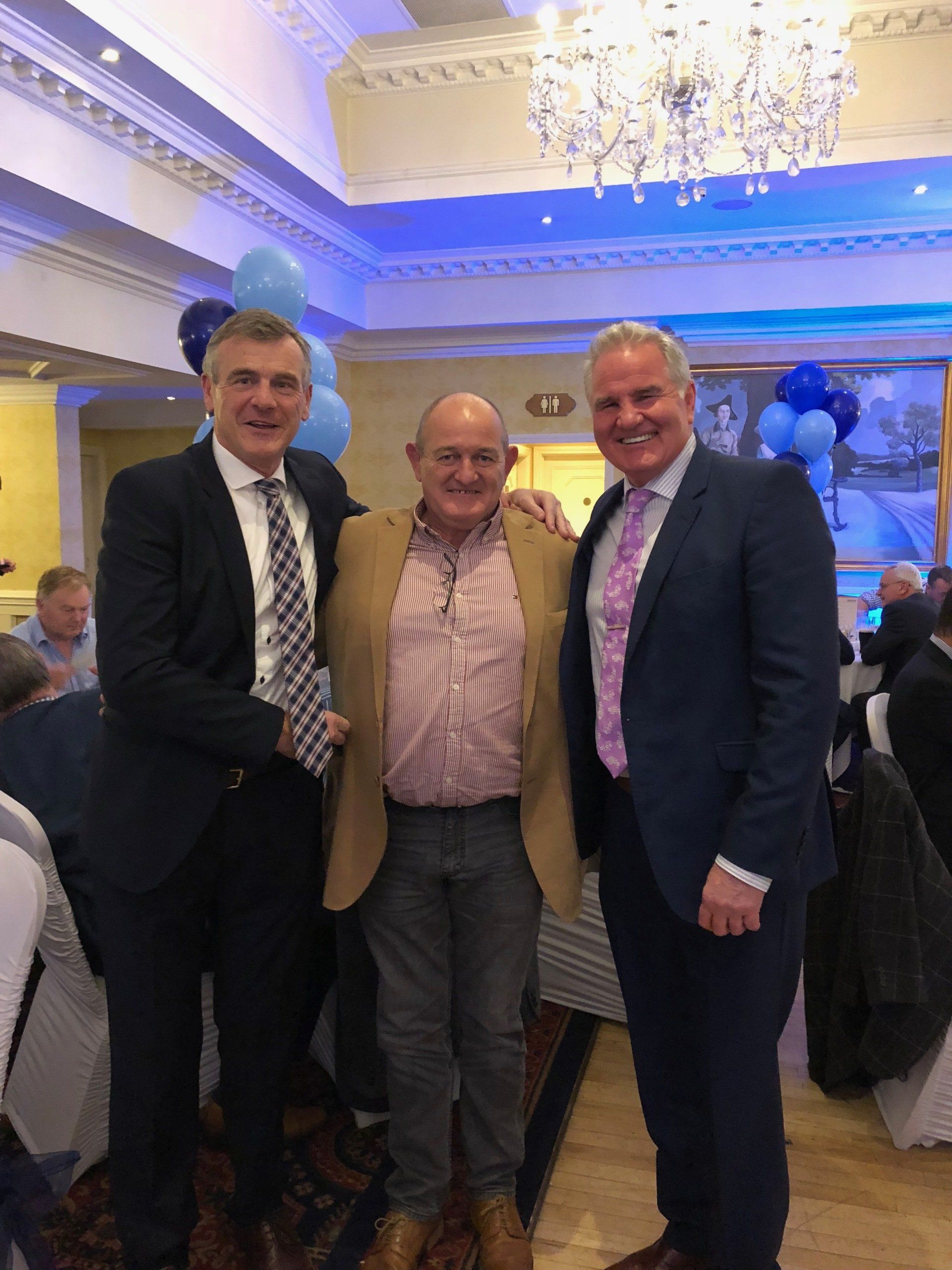 Three men smiling, arm around each other. Two in suits, one in a tan jacket, indoors with blue decor.