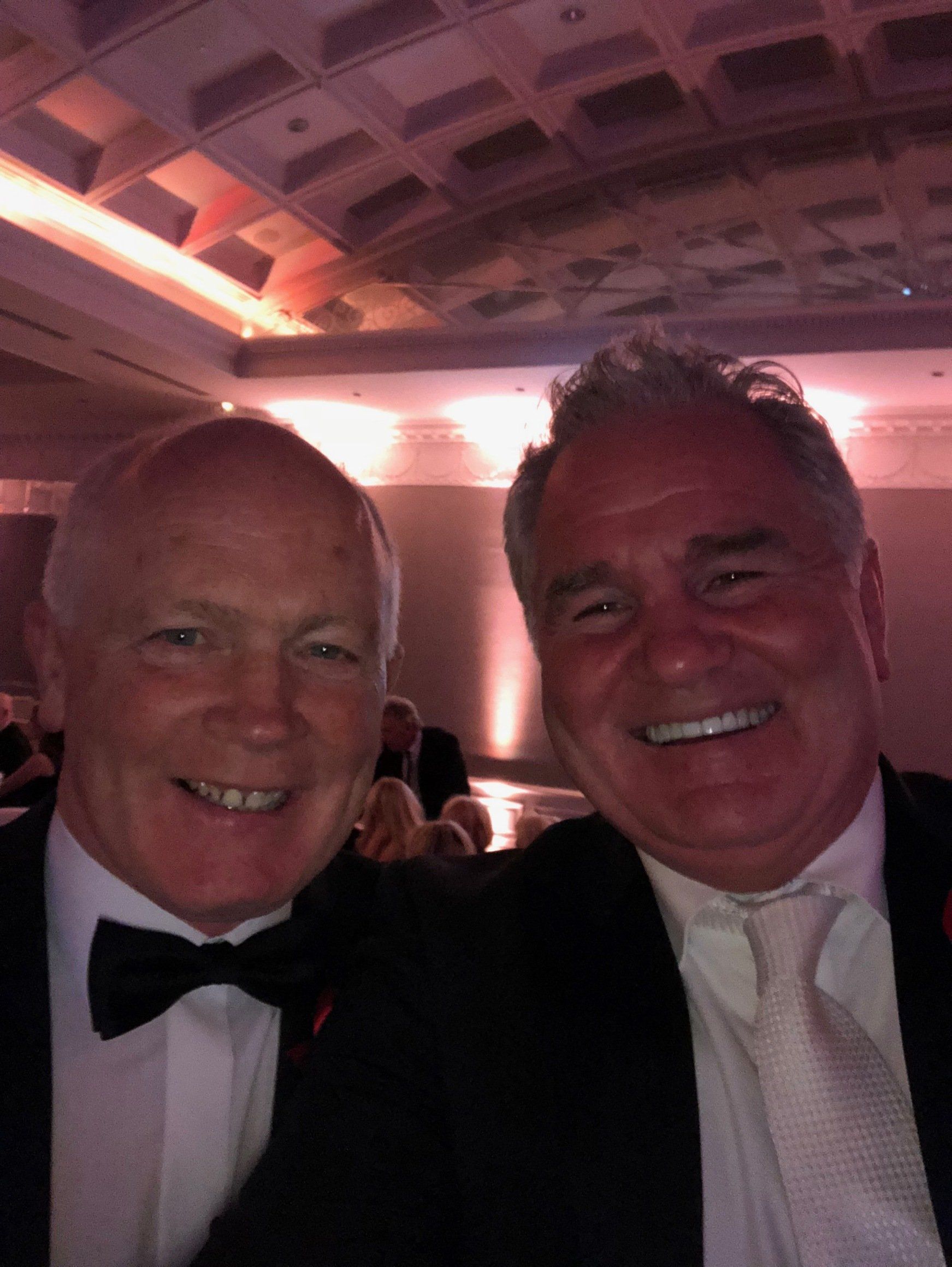 Two men in formal attire smiling at the camera at an event, likely a ballroom.