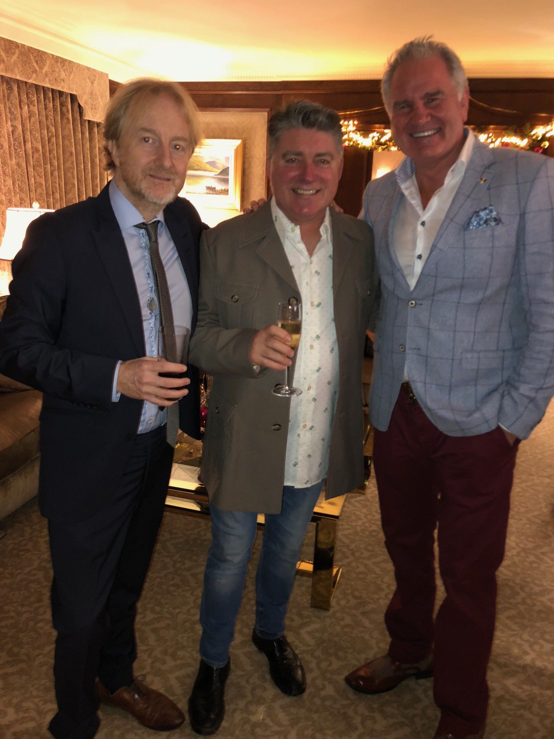 Three men smiling, indoors. Man in center holds champagne. One wears a suit, others jackets.