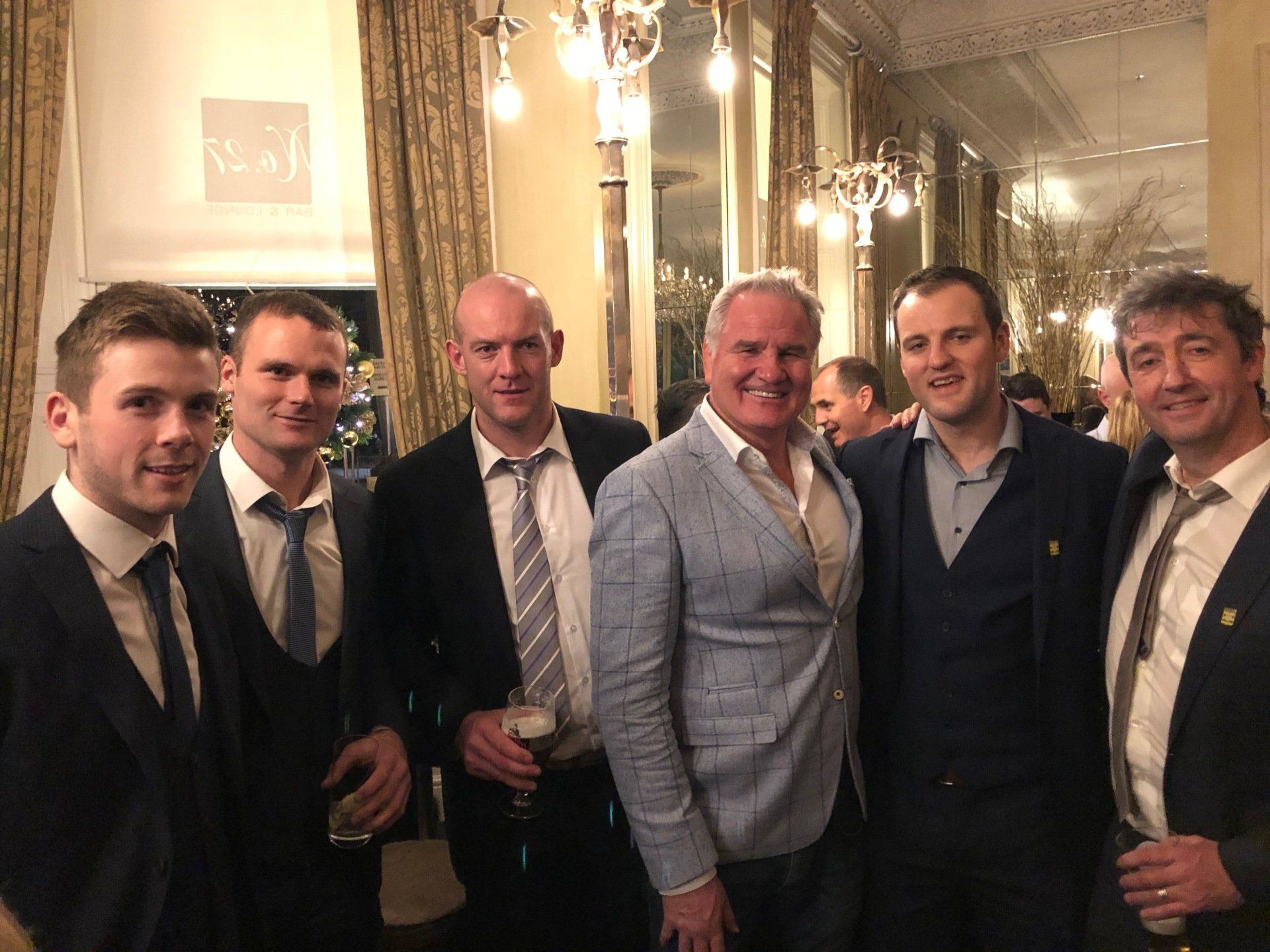 Group of men in suits, smiling in a well-lit room with chandeliers and mirrors.