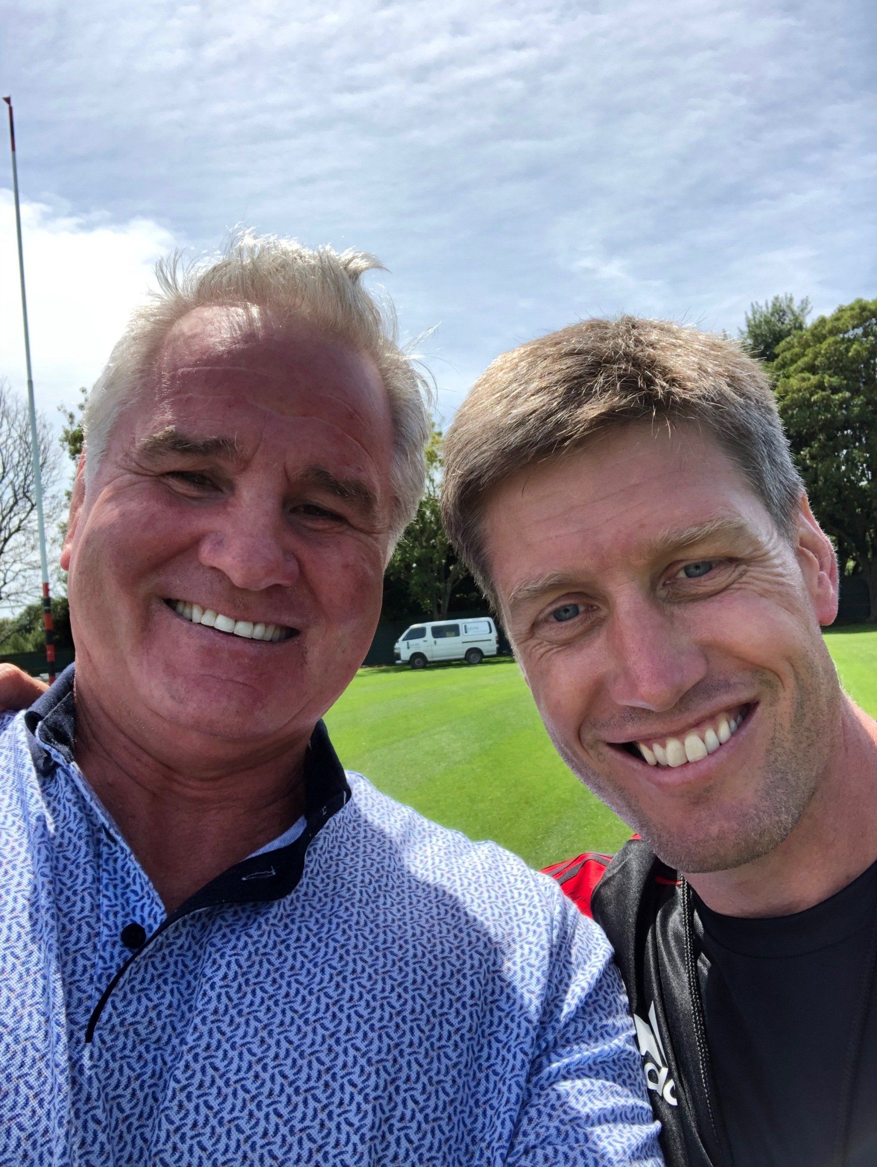 Two men smiling, taking a selfie outdoors on a sunny day. One in a blue patterned shirt, the other in a black shirt.