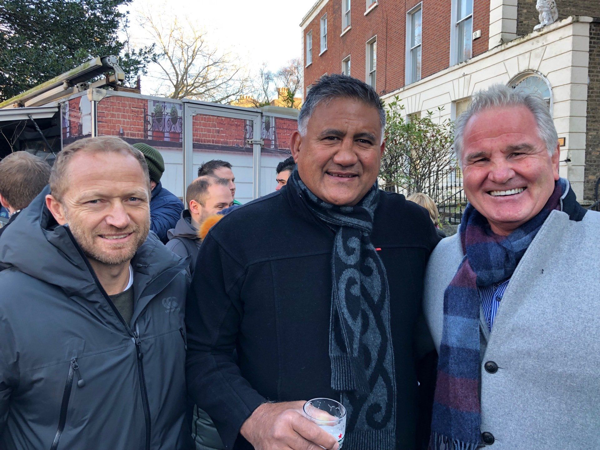 Three men smiling outdoors, posing for a photo. One holds a drink, buildings and people in background.