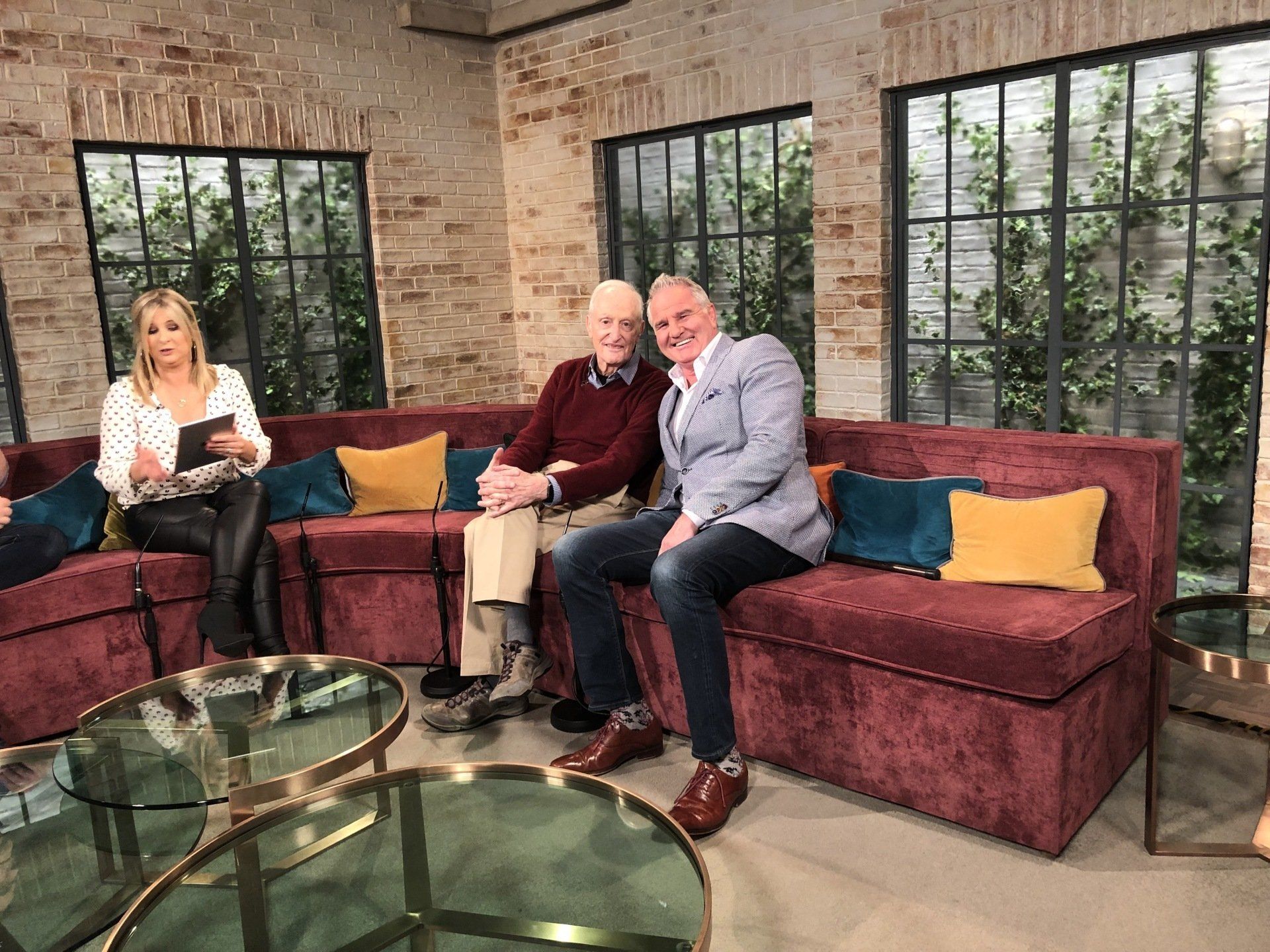 A TV studio with three people on a burgundy couch. Two men sit together, smiling. A woman reads notes.