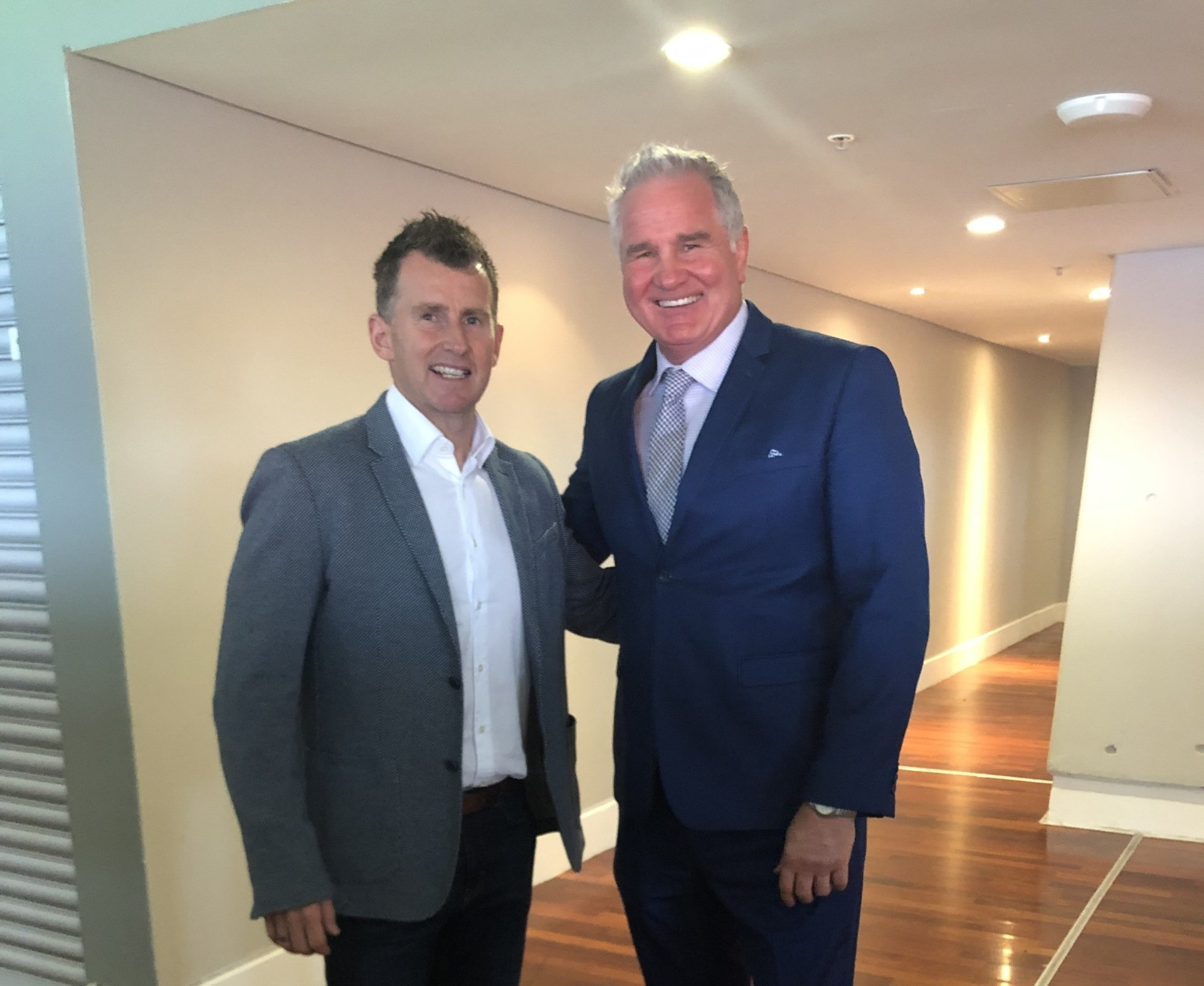 Two men standing together in a hallway. One in a blue suit, the other in a gray blazer.