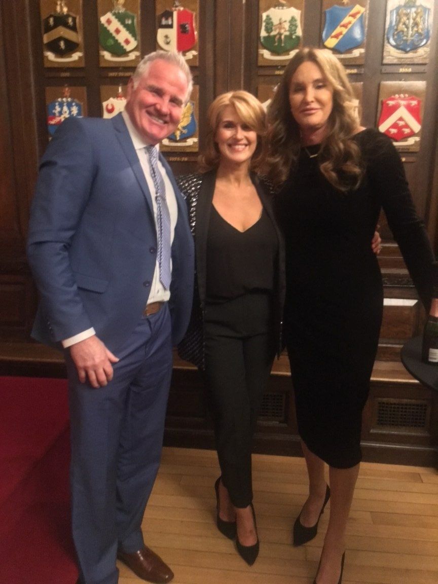 Three people smiling, posed together: a man in a blue suit, two women in black outfits, standing in front of crests.