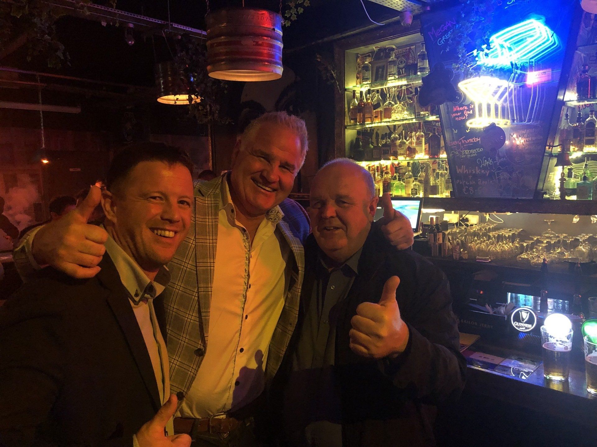 Three men smiling, giving thumbs up inside a bar. Background has bottles and neon signs.