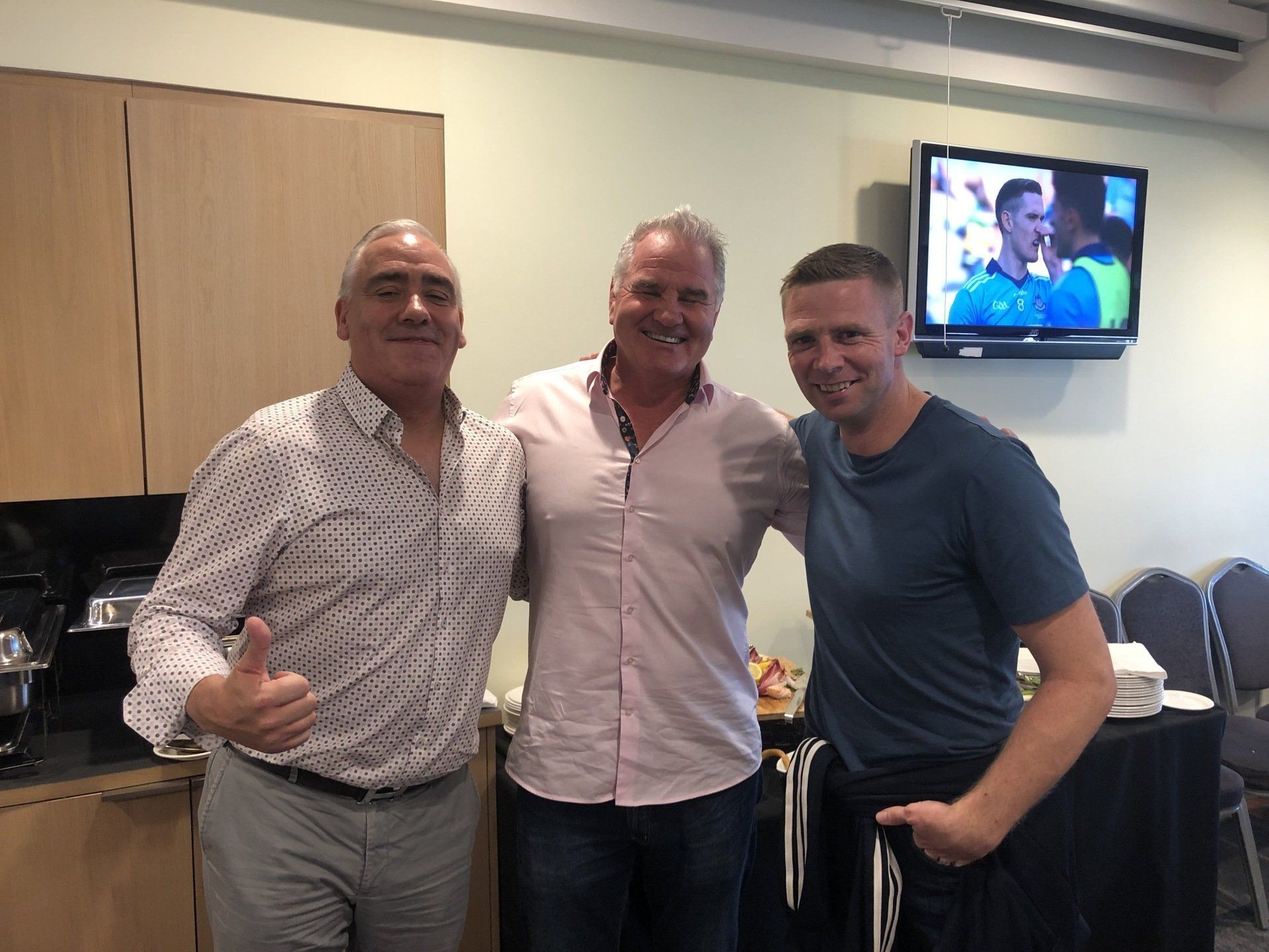 Three men pose together indoors. The man on the left gives a thumbs up. A TV shows a sporting event.