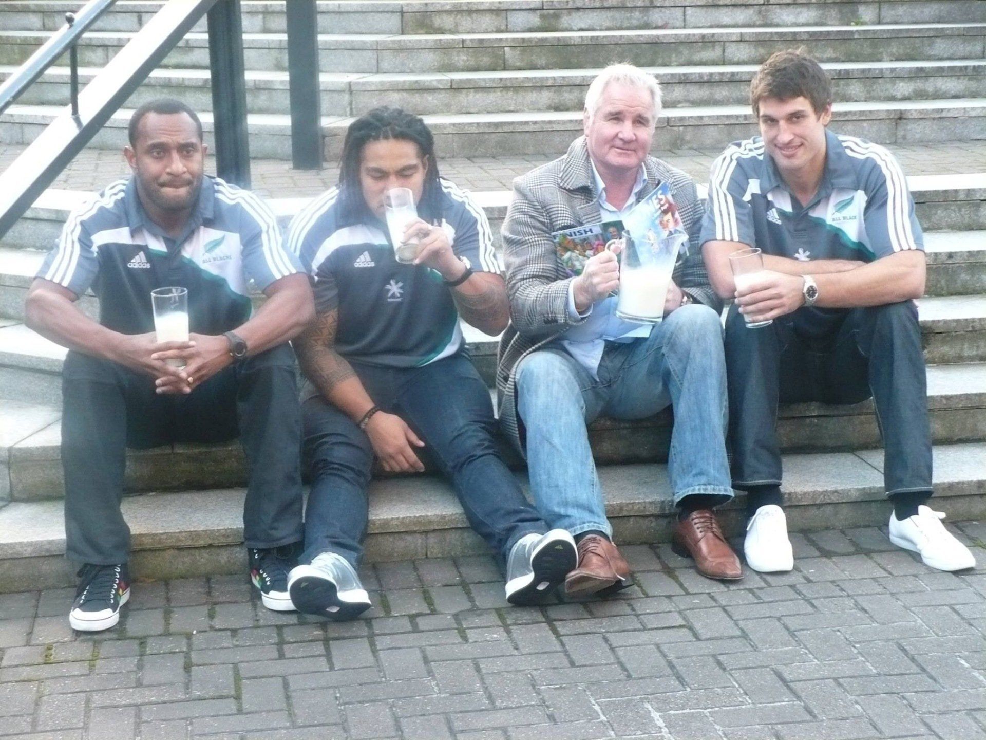 Four men sitting on steps, each holding a glass of milk. One is drinking. Gray and white clothing, brick steps.