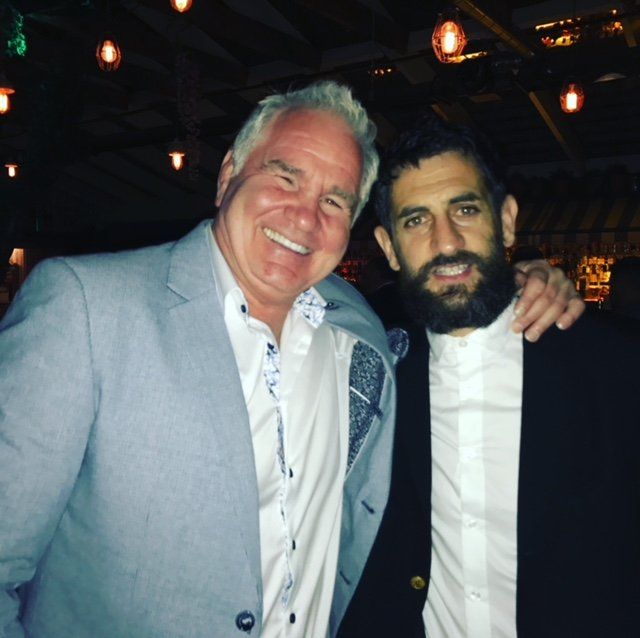 Two men smiling, one in a light blue suit, the other in a black suit, arm around the other.