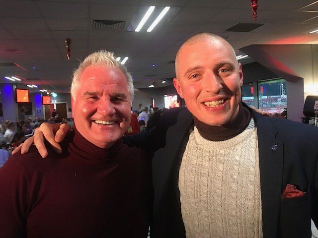 Two men smiling, one with white hair in a maroon turtleneck, the other bald in a navy blazer, both indoors.