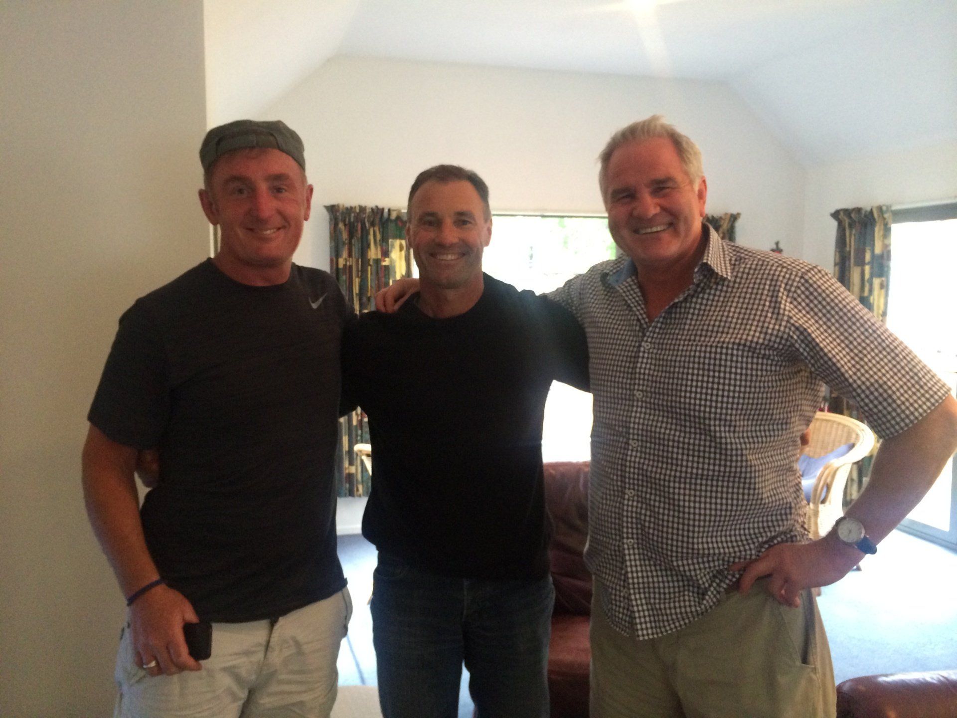 Three men standing indoors; one with arm around another's shoulder, all smiling.