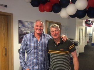 Two men smile, arm in arm, in a decorated room. One wears a plaid shirt, the other a striped polo. Balloons overhead.