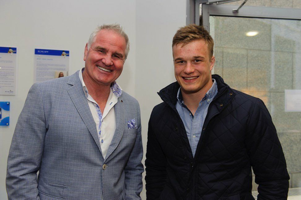 Two men smiling, one in a blue blazer and the other in a dark jacket, posing indoors near a window.